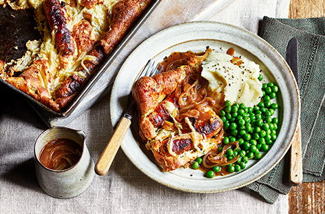 Put meaty sausages in a thick and fluffy Yorkshire pudding traybake and serve alongside mash, peas, and gravy